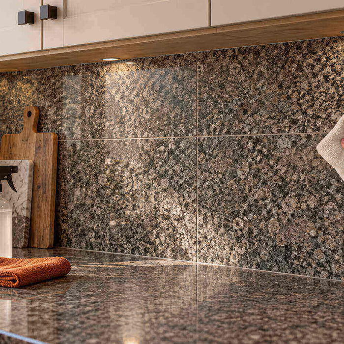 Hand wiping a gray granite tile backsplash with a soft cloth in a kitchen with white cabinets.