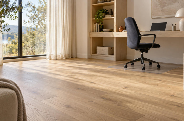 Bright home office with natural oak flooring, a built-in desk, and a dark ergonomic chair on a clear protective mat.
