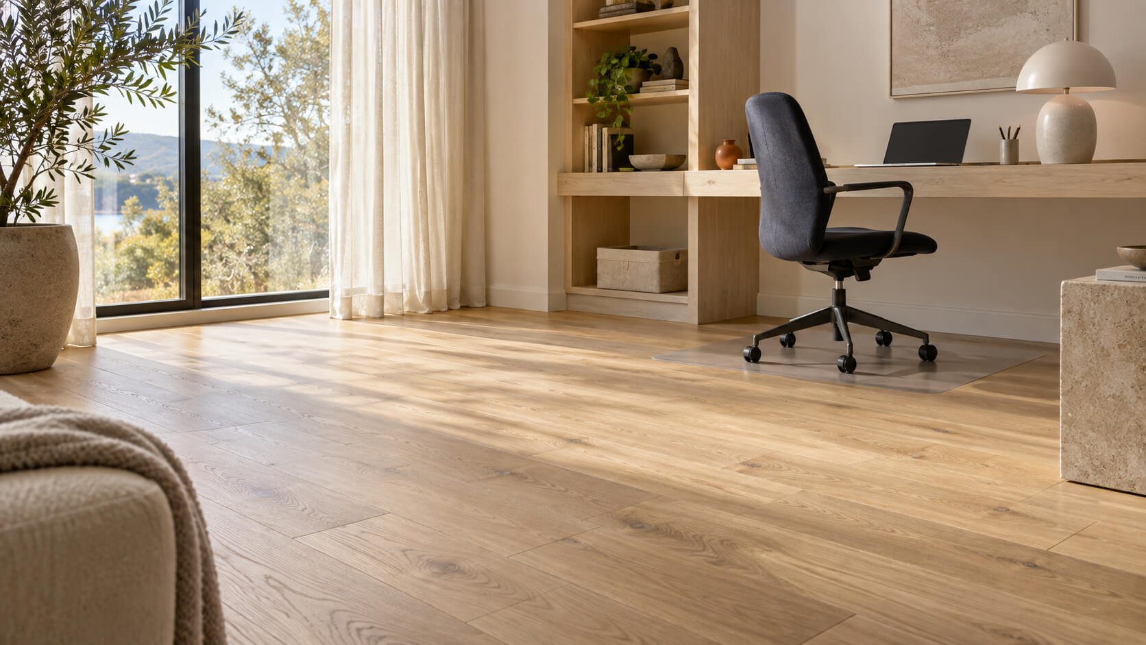 Bright home office with natural oak flooring, a built-in desk, and a dark ergonomic chair on a clear protective mat.