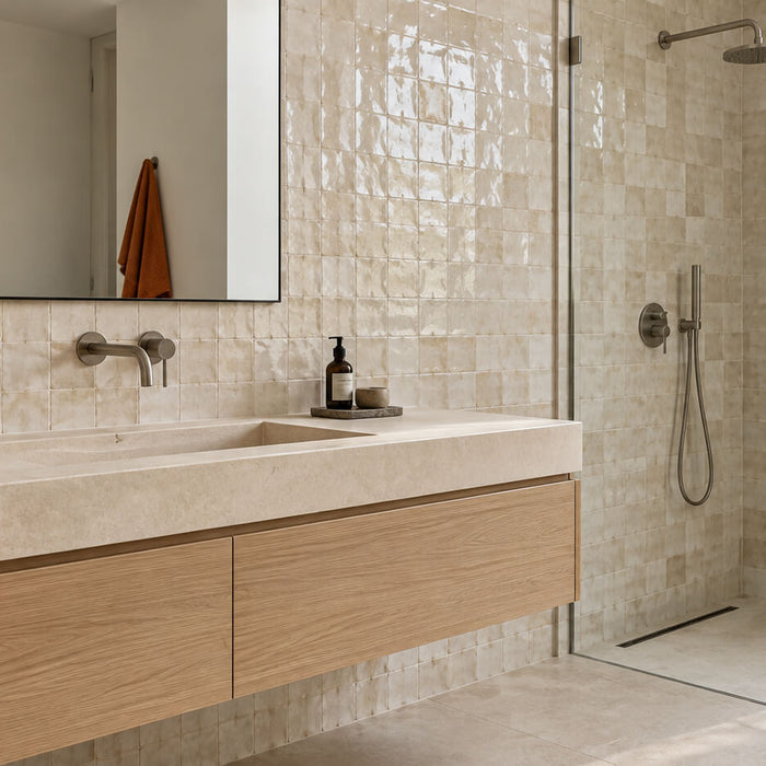 A warm, spa-like bathroom features glossy cream tiles, a floating oak vanity with an integrated stone sink, a large mirror, a glass walk-in rainfall shower, and earthy accents of terracotta, leafy branches, and burnt-orange towels in soft natural light.
