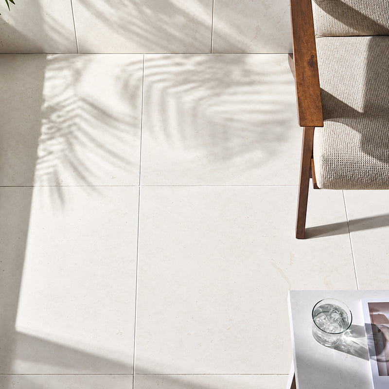 Corinthian White 24x24 filled and honed limestone flooring beside a wooden chair, with natural sunlight and soft leaf shadows.