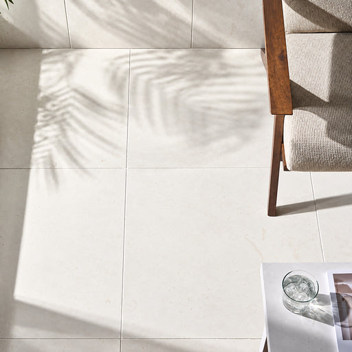 Corinthian White 24x24 filled and honed limestone flooring beside a wooden chair, with natural sunlight and soft leaf shadows.