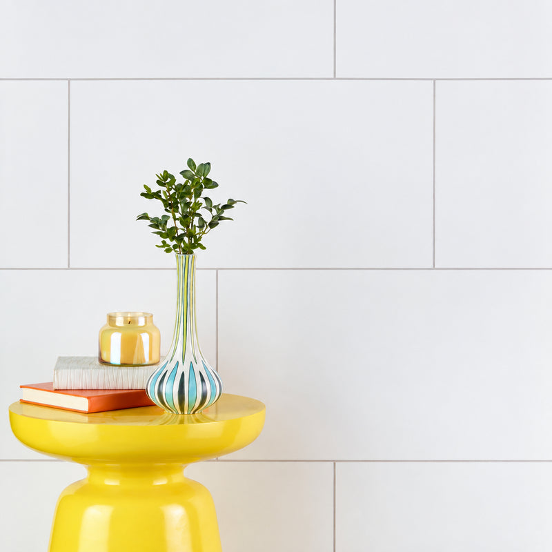 Iceland White porcelain wall tiles behind a yellow side table with books, a candle and a striped vase.