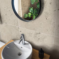 Jura Grey honed limestone bathroom wall tiles behind a white vessel sink and a round black-framed mirror.
